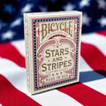 Bicycle Stars and Stripes Playing Cards sealed deck featuring patriotic red, white and blue artwork, photographed against an American flag background.