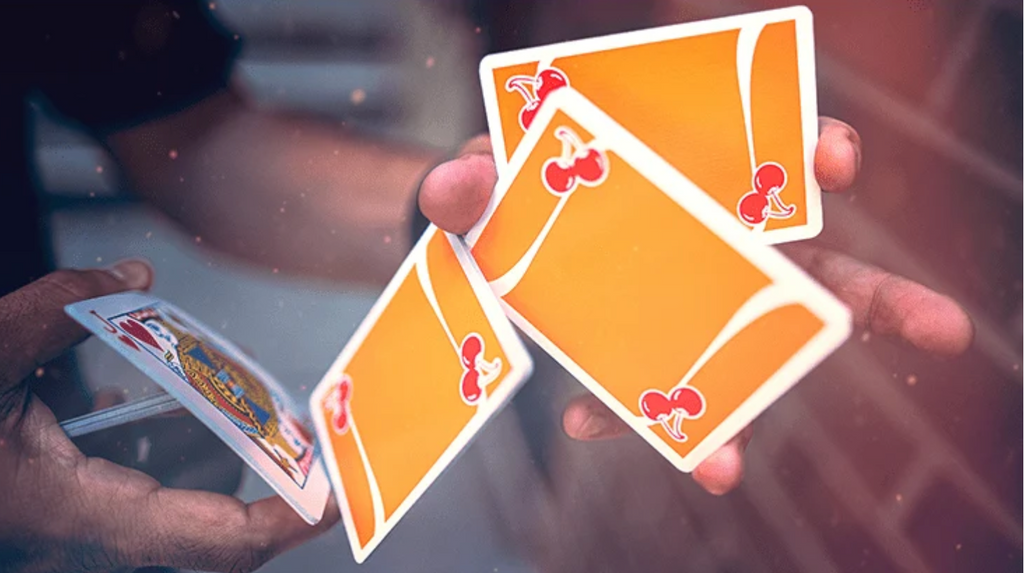 Close-up of Cherry Casino Summerlin Sunset Orange Playing Cards during a cardistry flourish, showing vibrant orange card backs with red cherry artwork, flowing white stripes and white borders.