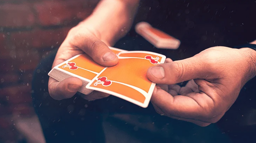 Close-up of Cherry Casino Summerlin Sunset Orange Playing Cards being handled during a card flourish, showing the vibrant orange card backs, red cherry artwork and flowing white stripes.