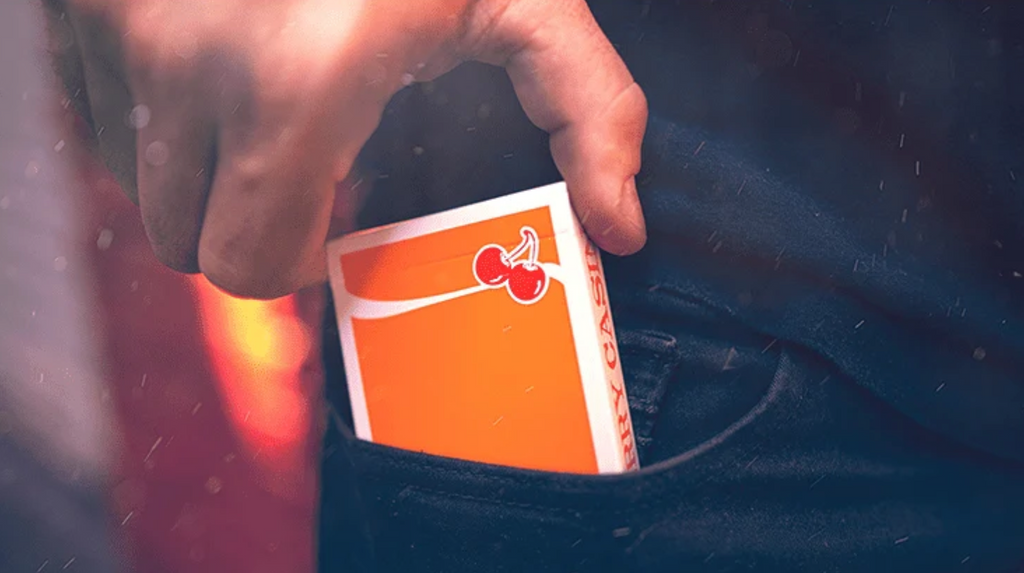 Cherry Casino Summerlin Sunset Orange Playing Cards being placed into a jeans pocket, showing the vibrant orange tuck box, red cherry artwork and white casino-inspired design.