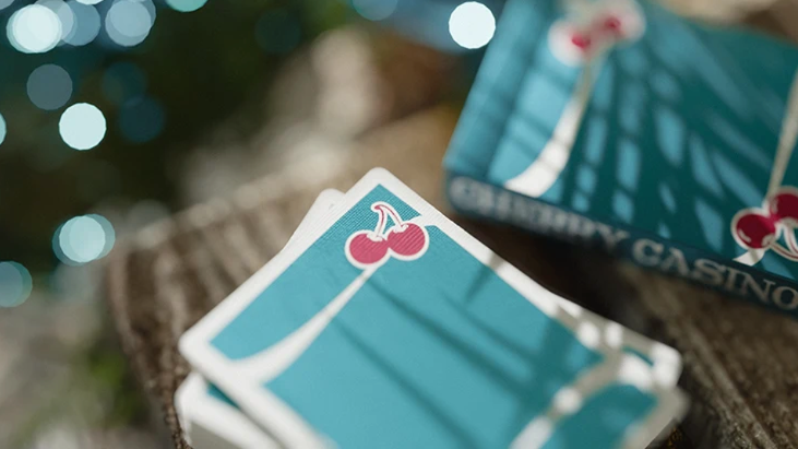 Close-up of Cherry Casino Tropicana Teal Playing Cards showing the turquoise teal card backs with red cherry artwork and flowing white stripes, alongside the matching tuck box.