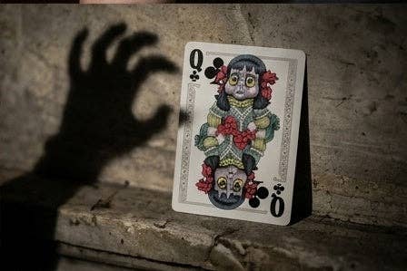 Haunted Dolls Playing Cards by CPC featuring the custom Queen of Clubs with eerie porcelain doll artwork, displayed against an aged stone wall with a sinister reaching hand shadow.