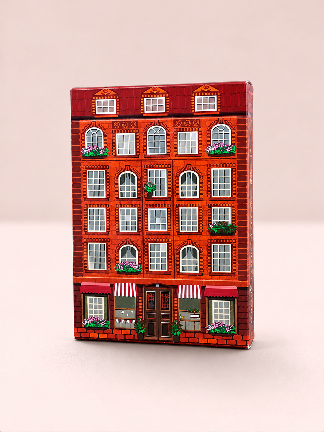 Front view of the House Playing Cards tuck box, designed as a detailed red-brick townhouse with white windows, flower boxes, striped awnings and a central doorway