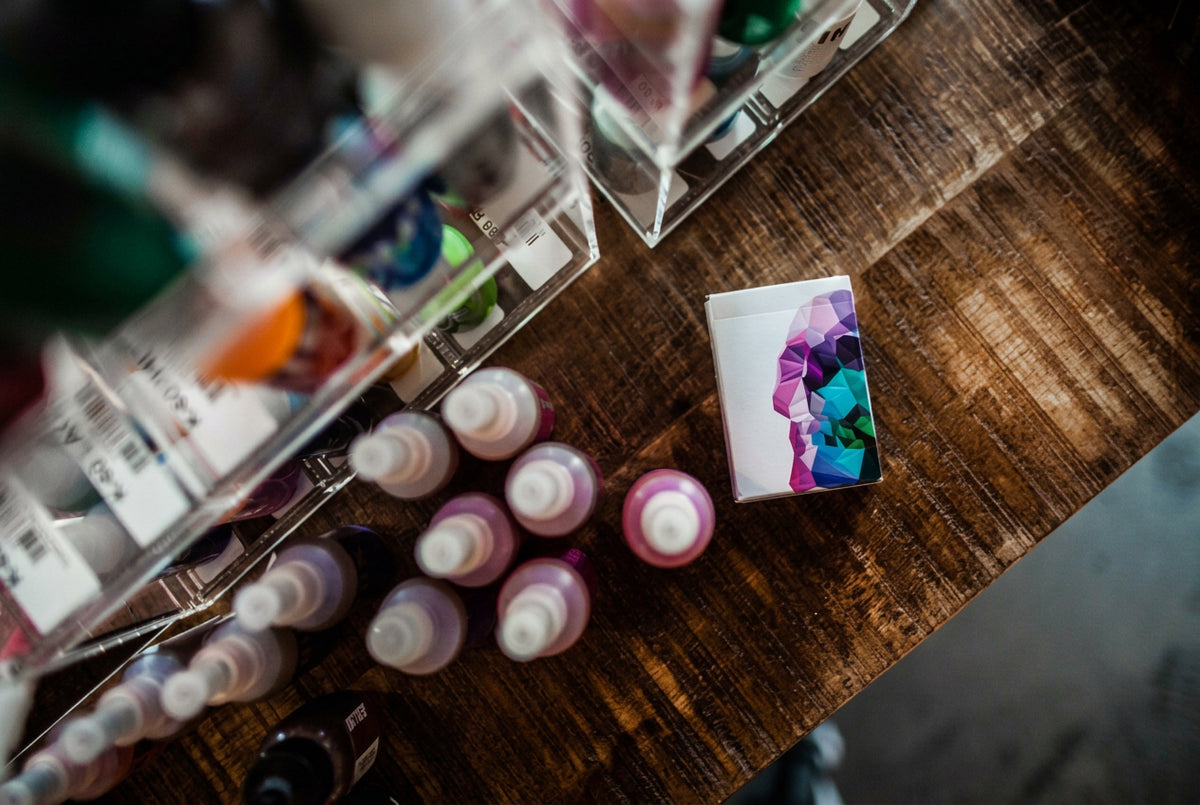 Memento Mori Playing Cards by Chris Ramsay – Original deck featuring colourful geometric skull artwork, photographed on a wooden table beside art supplies.