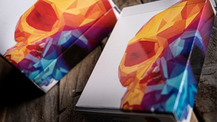 Two Memento Mori Genesis Playing Card tuck boxes featuring vibrant geometric skull artwork in orange, red, purple and blue, photographed on a wooden surface.