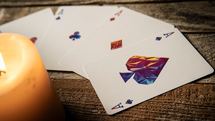 Memento Mori Genesis Playing Cards showing four custom Aces with colourful geometric suit designs, displayed on a rustic wooden surface beside a lit candle.