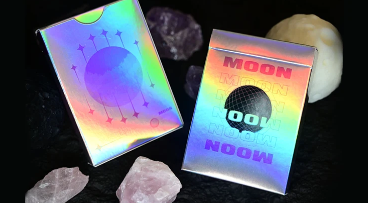 Moon White Holo Playing Cards by Hiddenlight showing the front and back of the iridescent holographic tuck box, featuring rainbow MOON lettering, a black wireframe sphere and colourful lunar artwork.