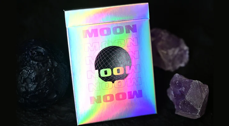 Moon White Holo Playing Cards by Hiddenlight featuring an iridescent rainbow holographic tuck box with colourful MOON lettering and a black wireframe lunar sphere, displayed alongside dark lunar rocks and purple crystals.