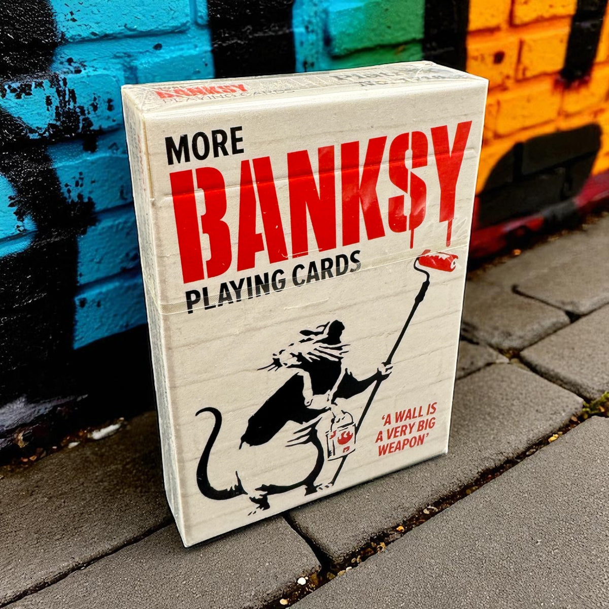 Piatnik More Banksy playing card box beside a colourful painted brick wall