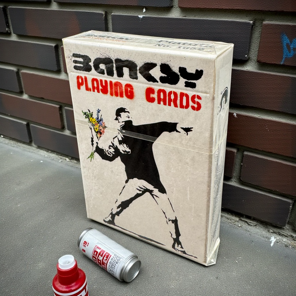 Piatnik Banksy First Collection playing card box featuring flower thrower artwork beside a brick wall and spray-paint cans