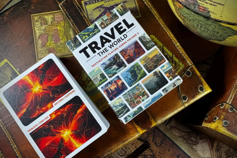 Travel the World Volume 2 playing cards deck displayed on vintage travel maps and suitcases, featuring a volcanic lightning playing card and colourful destination illustrations.